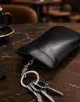 A black leather key pouch holding silver keys, placed on a dark marble table in a cafe setting with a coffee cup and candle in the background.