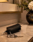 A black pebbled leather key pouch with a silver key ring and clasp, resting on a marble countertop in a warm, modern interior setting.