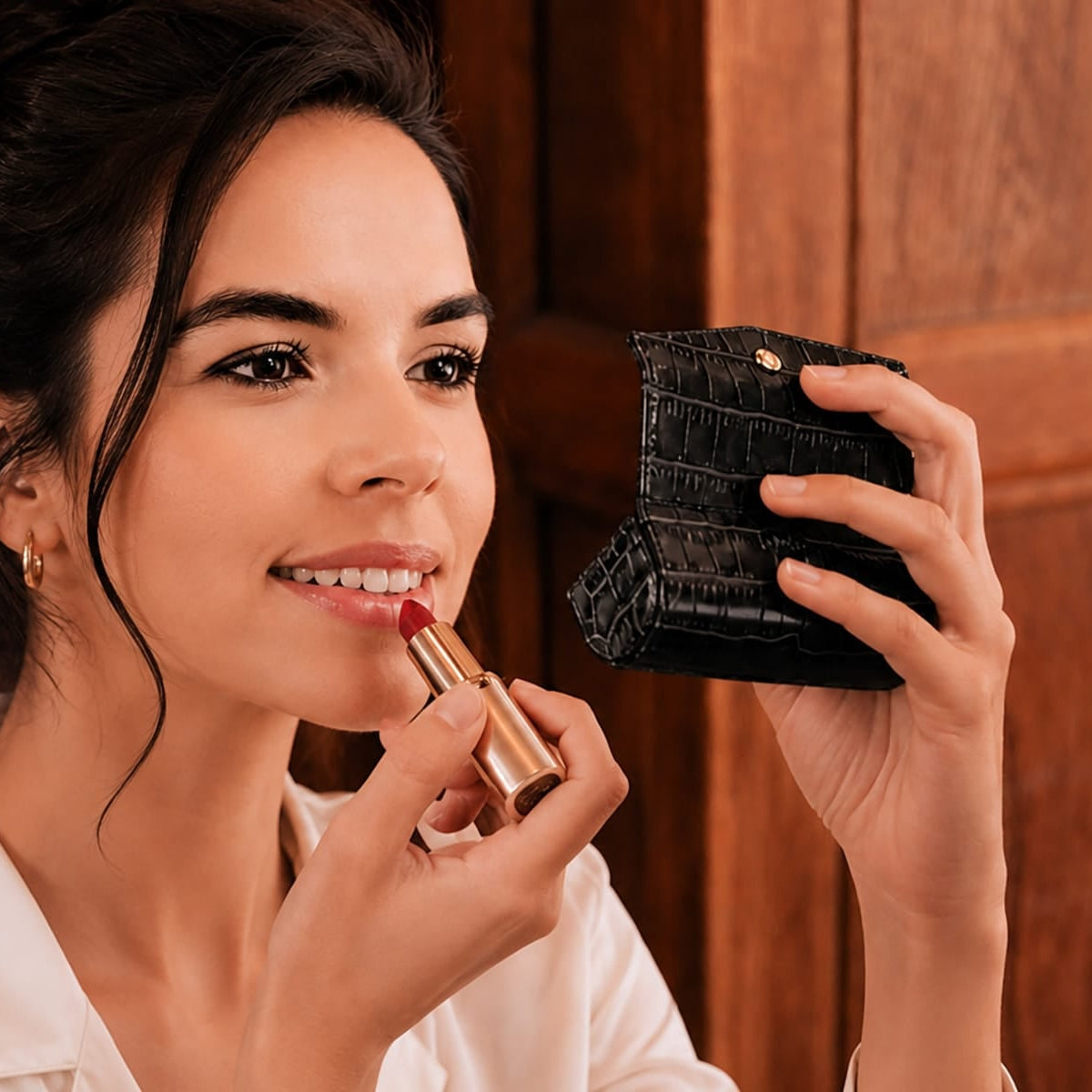 A woman with dark hair in an updo applies a red lipstick while looking into a black crocodile-embossed compact mirror. She is wearing a white collared shirt against a warm brown wooden background.