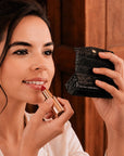 A woman with dark hair in an updo applies a red lipstick while looking into a black crocodile-embossed compact mirror. She is wearing a white collared shirt against a warm brown wooden background.