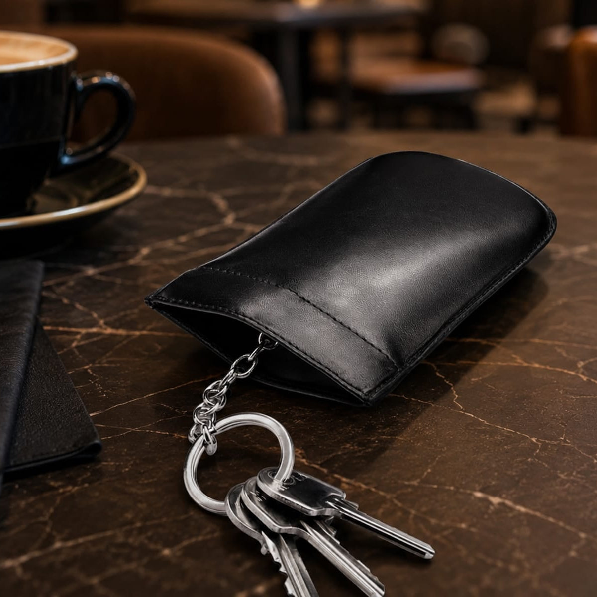 A black leather key pouch holding silver keys, placed on a dark marble table in a cafe setting with a coffee cup and candle in the background.