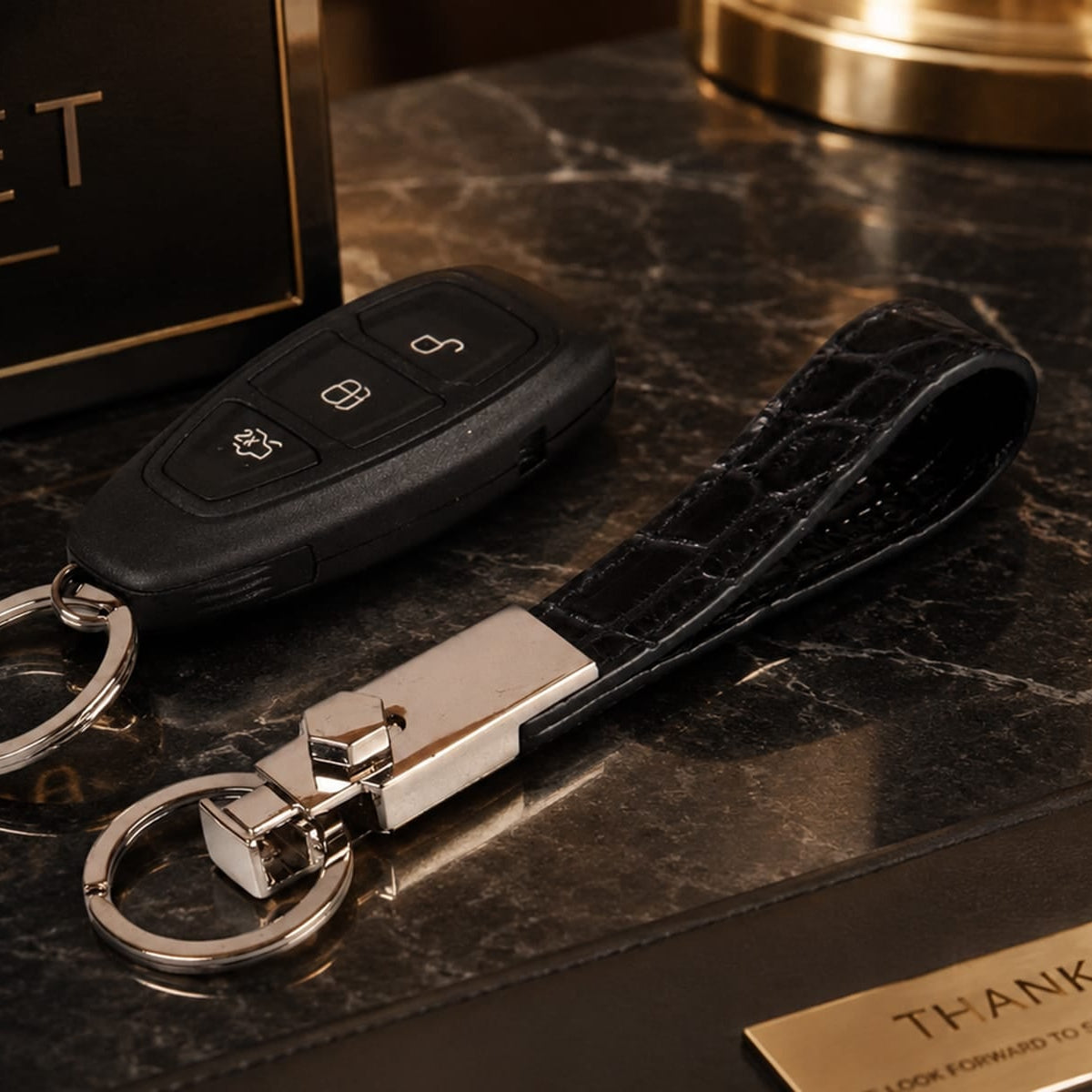 A luxury keychain set featuring a black car key fob, a black crocodile-embossed leather wristlet strap, and a polished silver-toned metal clasp, displayed on a dark marble surface next to a branded black box.