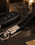 A luxury keychain set featuring a black car key fob, a black crocodile-embossed leather wristlet strap, and a polished silver-toned metal clasp, displayed on a dark marble surface next to a branded black box.