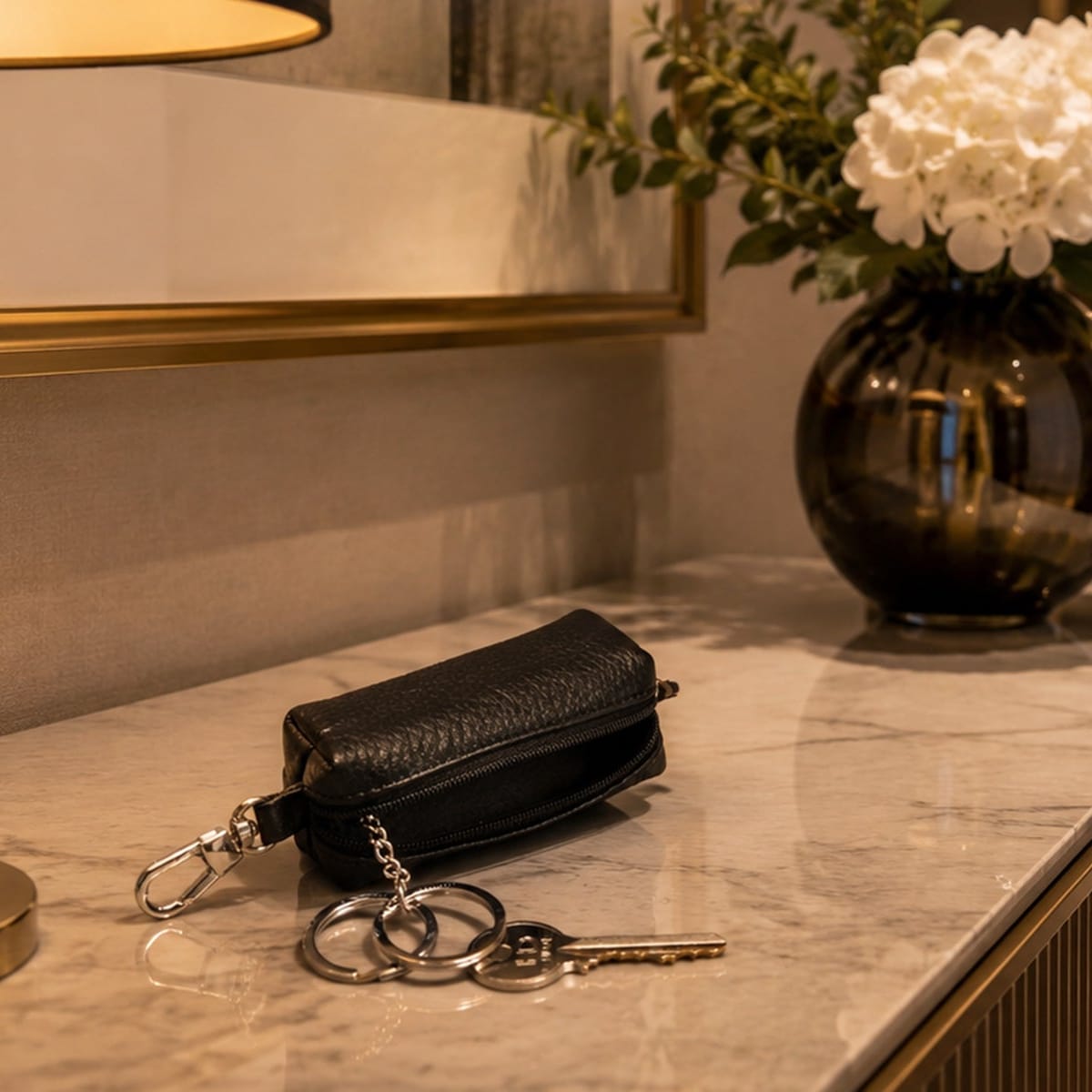 A black pebbled leather key pouch with a silver key ring and clasp, resting on a marble countertop in a warm, modern interior setting.